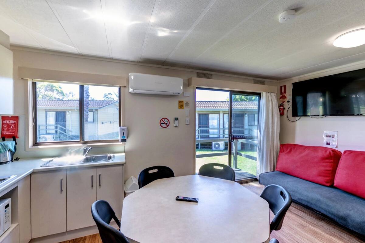 A compact kitchenette with a stove, microwave, and sink, a round table with chairs, a red couch, and a TV mounted on the wall.