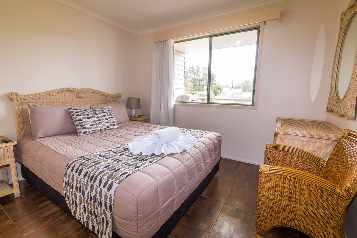 A neatly made bed with a patterned bedspread, a bedside lamp, a wicker chair, and a window with curtains.