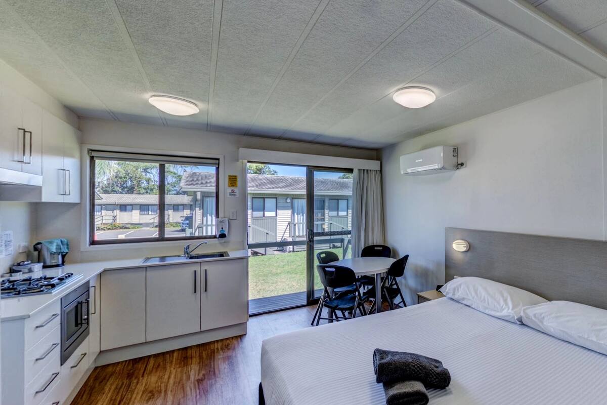 A modern hotel room with a large bed, a kitchenette, a dining area, and a sliding glass door to the outside.