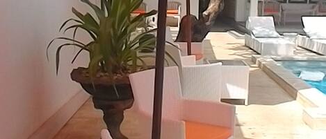 A poolside area with white wicker chairs and orange cushions, a large orange umbrella, and a potted plant.
