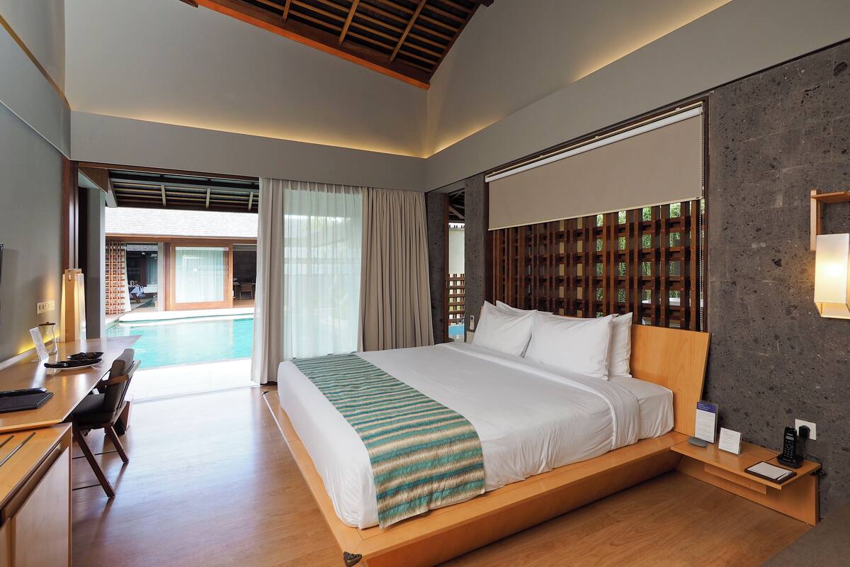 A modern hotel room with a large bed, wooden headboard, a desk with a chair, a small table, and a view of an outdoor pool area.