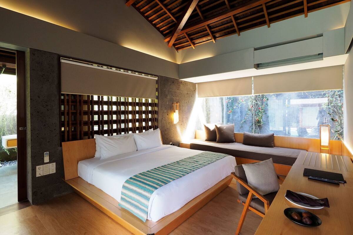 A modern hotel room with a bed, seating area, and large windows.