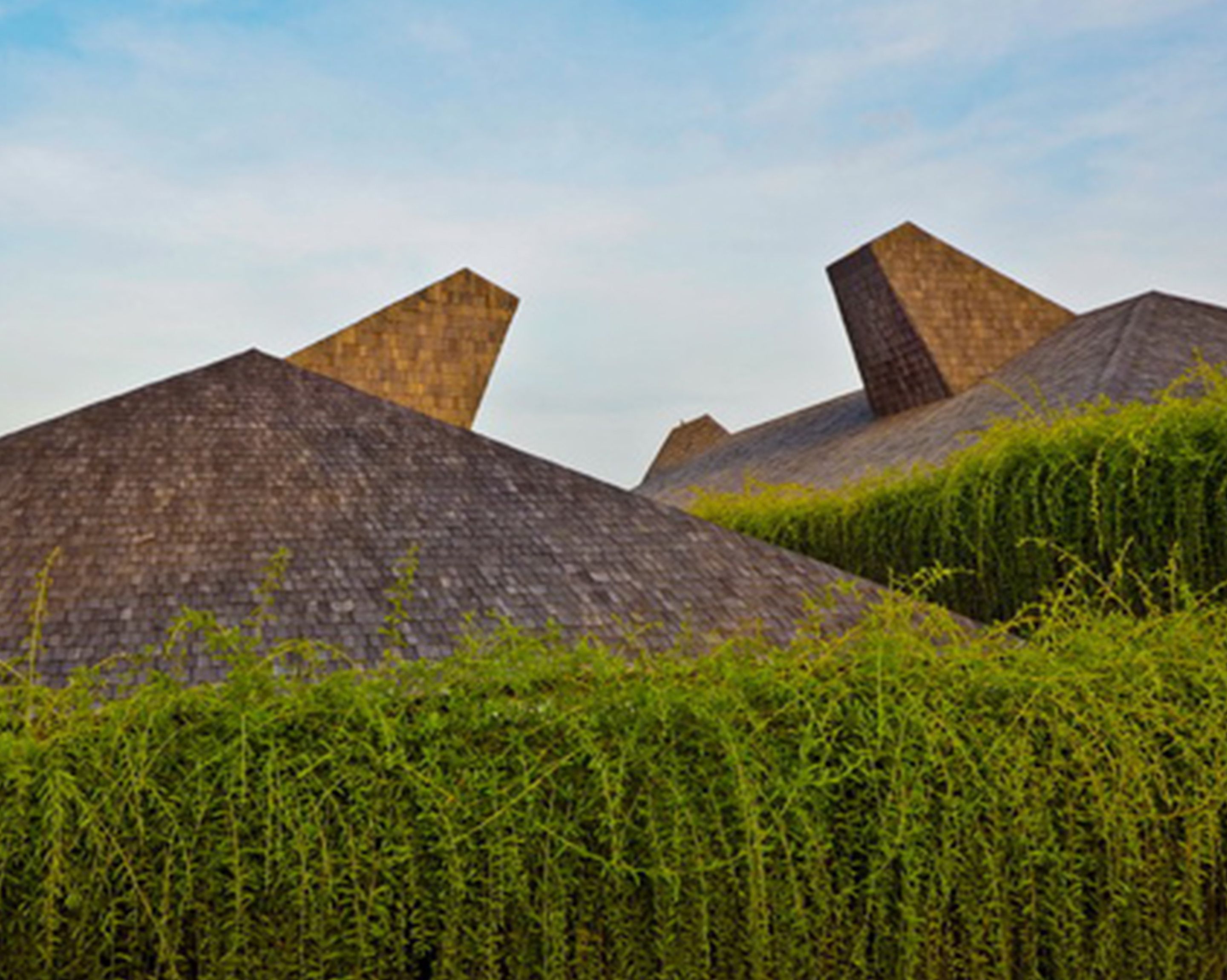 Conjunto de edifícios modernos com design geométrico e telhados inclinados, cercado por vegetação densa e exuberante.