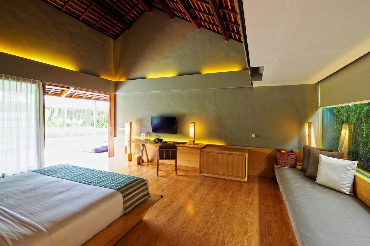A hotel room with a large bed, a sofa, a desk with a flat-screen TV, and a large window with a view of greenery.