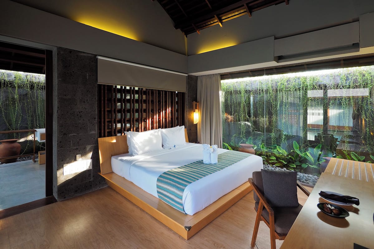 A modern hotel room with a large bed, a wooden headboard, a dining table, and a view of greenery through large windows.