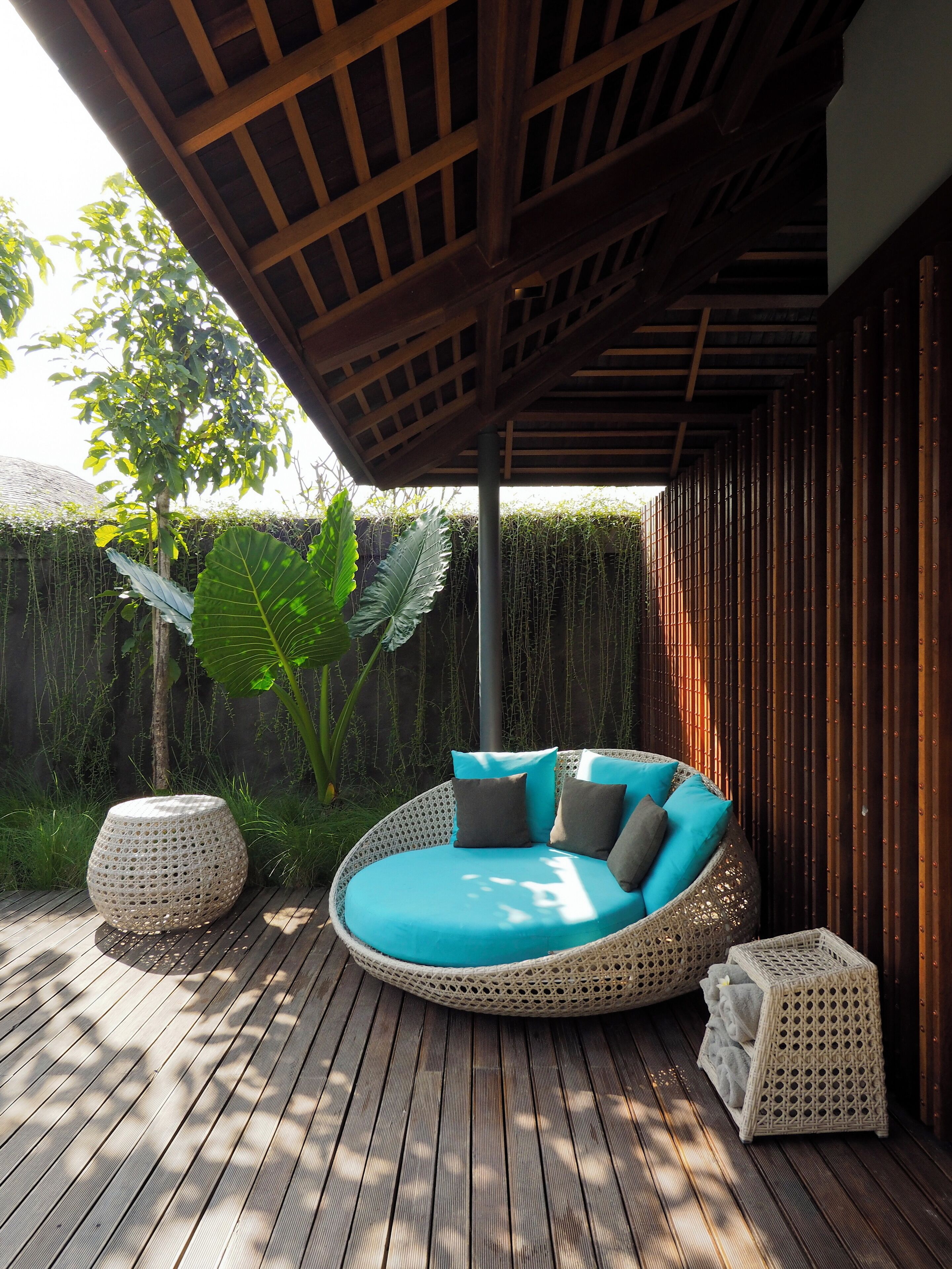 A round wicker sofa with cushions, a small round table, and a storage chest on a wooden deck.