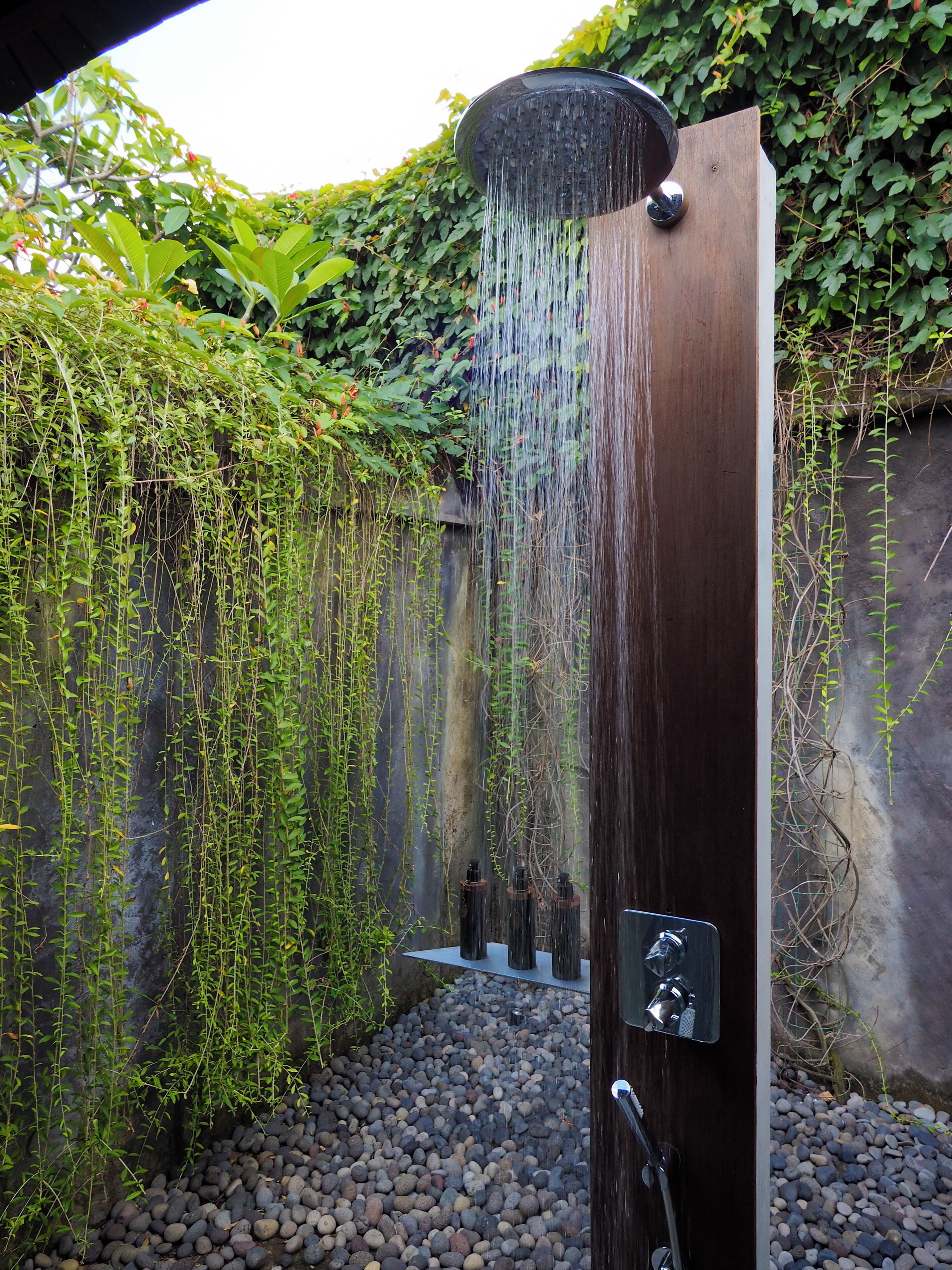 Una ducha exterior moderna con una fuente de agua vertical, rodeada de follaje y un suelo de guijarros.