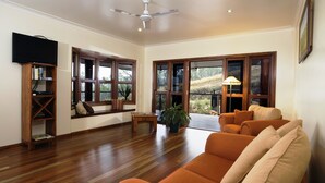 Un salón espacioso con piso de madera, un sofá grande color naranja, un televisor montado en un estante de madera y ventanas amplias con vistas al exterior.