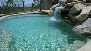 Piscina cristalina con caída de agua, rodeada por una terraza de hormigón y una valla metálica.