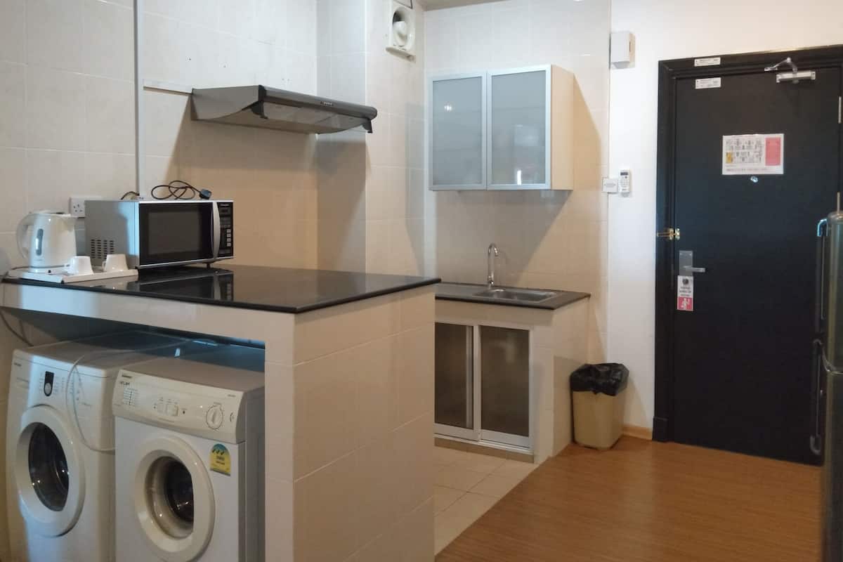 Een kleine keuken met een magnetron, waterkoker en wasmachine, een spoelbak, en een zwarte deur met een bericht.