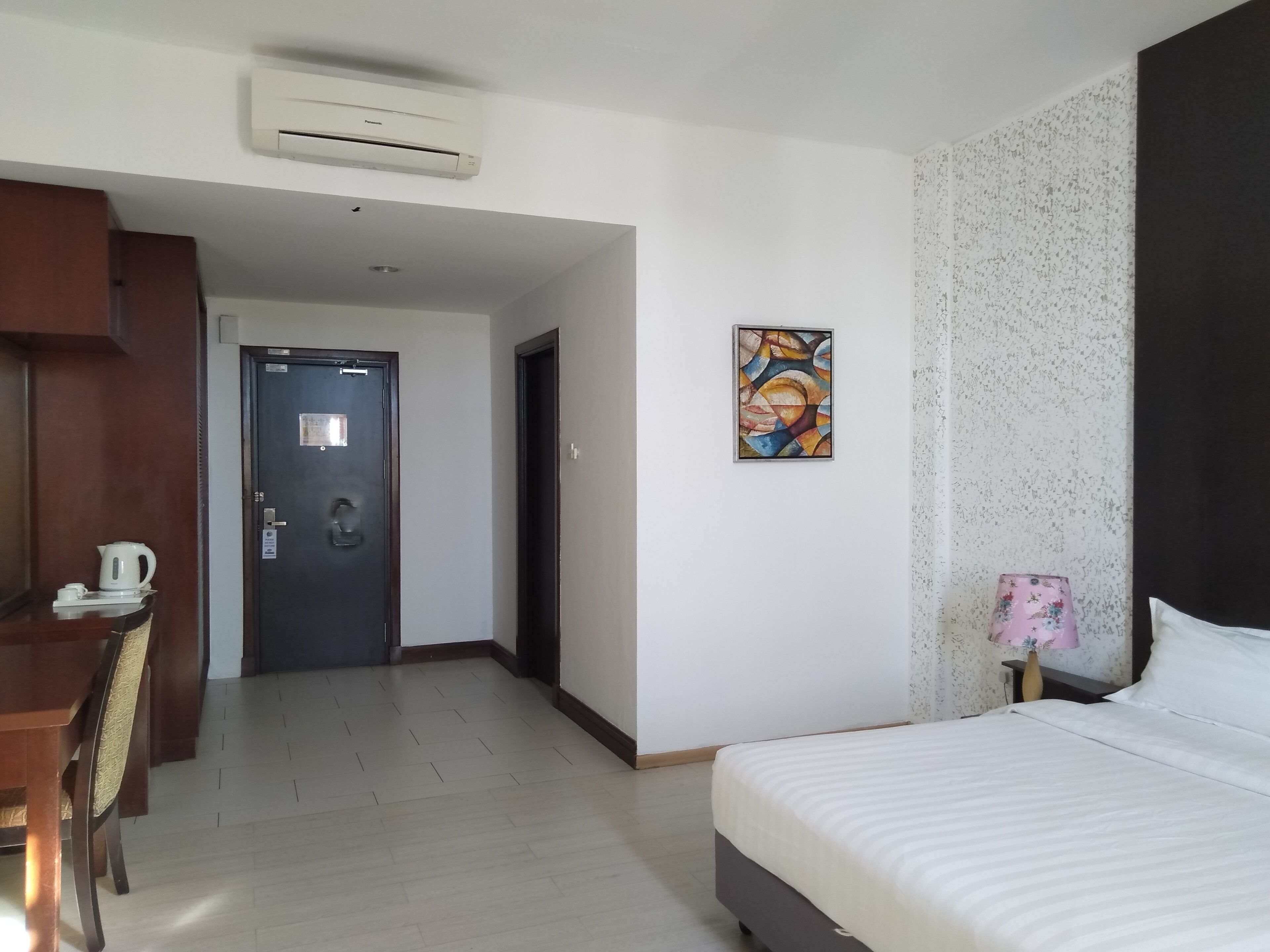 Quarto de hotel com cama, escrivaninha, cadeira, porta e ar-condicionado fixado na parede.