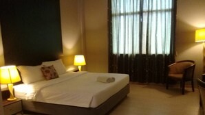 Una cama bien tendida con sábanas blancas, dos lámparas de noche, una silla y una ventana con cortinas.