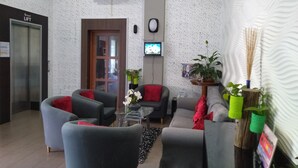 Un vestíbulo con sillones tapizados en gris y rojo, una mesa de centro, una planta en maceta, un televisor montado en la pared y una puerta con la etiqueta 'Ground LIFT'.