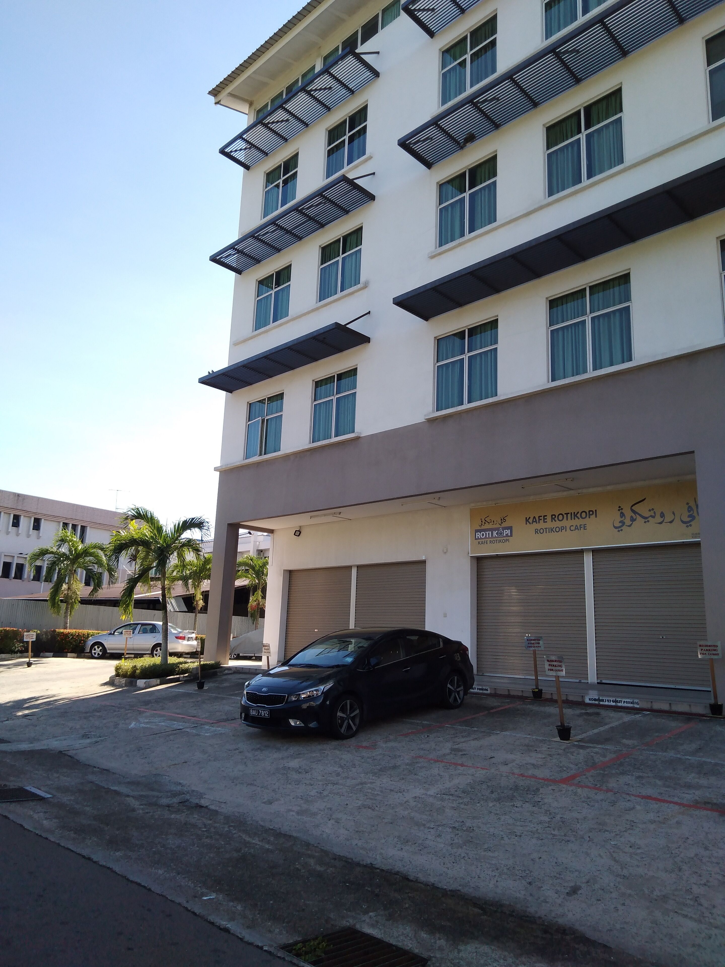 Un edificio de varios pisos con una cafetería llamada KAFE ROTIKOPI. Hay palmeras en frente, y un coche negro estacionado delante del edificio.