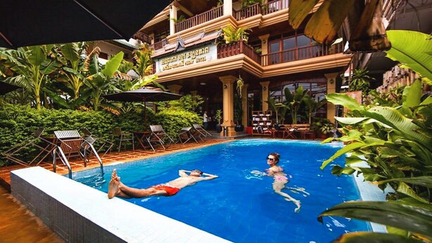 A swimming pool with two people in it, surrounded by lush greenery and a building with a sign that reads "Hotel". - Angkor Vattanak Pheap Hotel (Siem Reap)