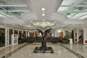 Un lobby espacioso con una gran araña central, una estatua alada imponente y un área de descanso con sofás y una mesita de centro.