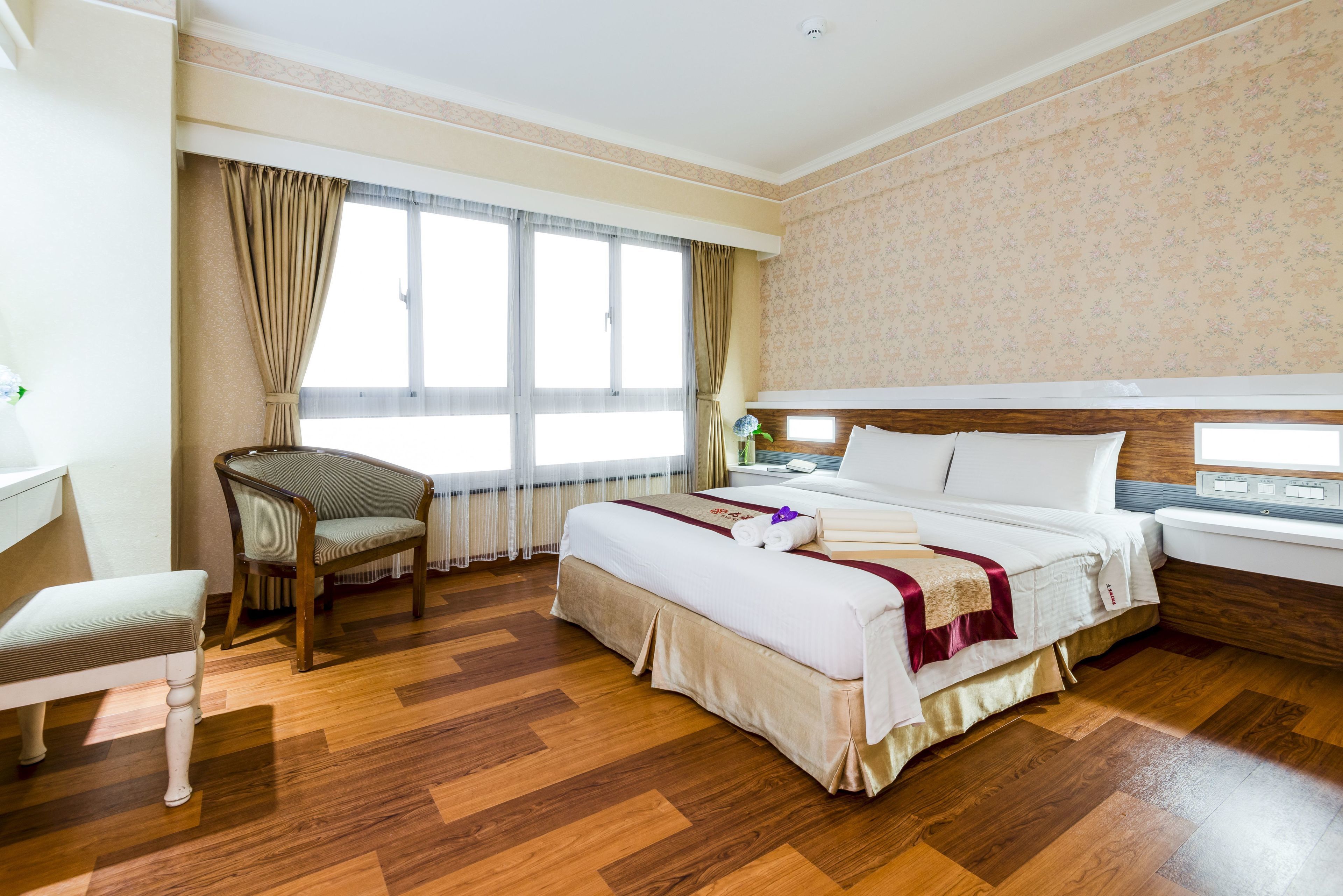 Ein Hotelzimmer mit einem großen Bett, einem Sessel, einem Schreibtisch und einem Fenster mit Vorhängen.