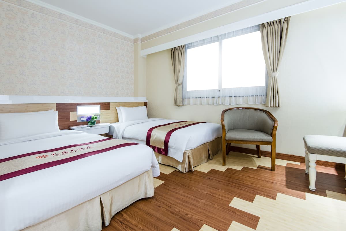 Hotellihuone, jossa on kaksi sänkyä, puinen tuoli, pieni pöytä maljakolla ja ikkuna, jossa on verhot.