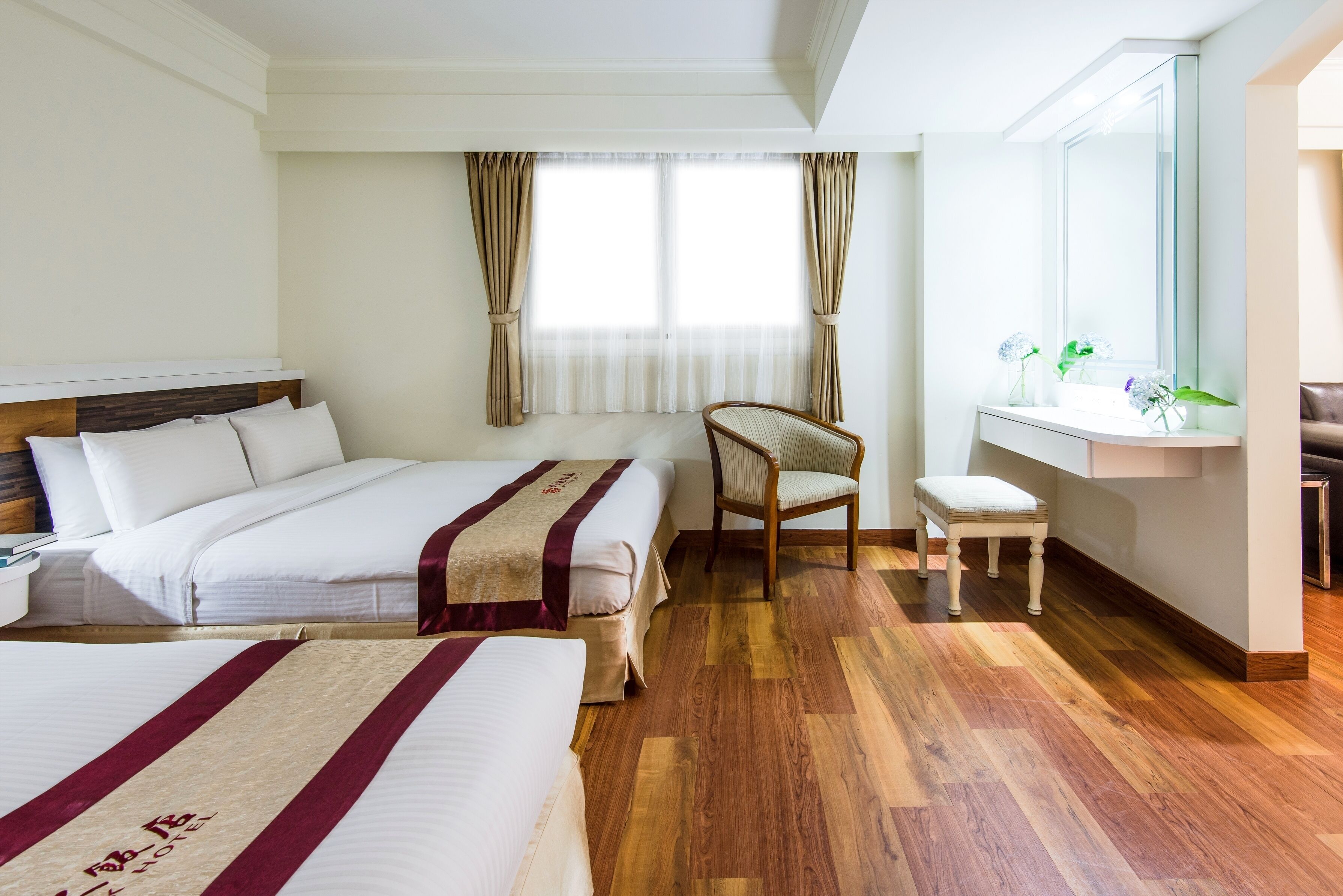 A hotel room with two beds, a wooden floor, a chair, a small table, and a window with curtains.
