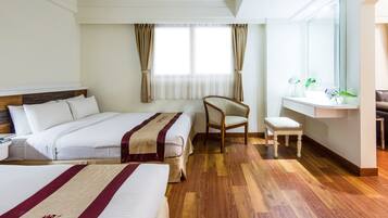 A hotel room with two beds, a wooden floor, a chair, a small table, and a window with curtains.
