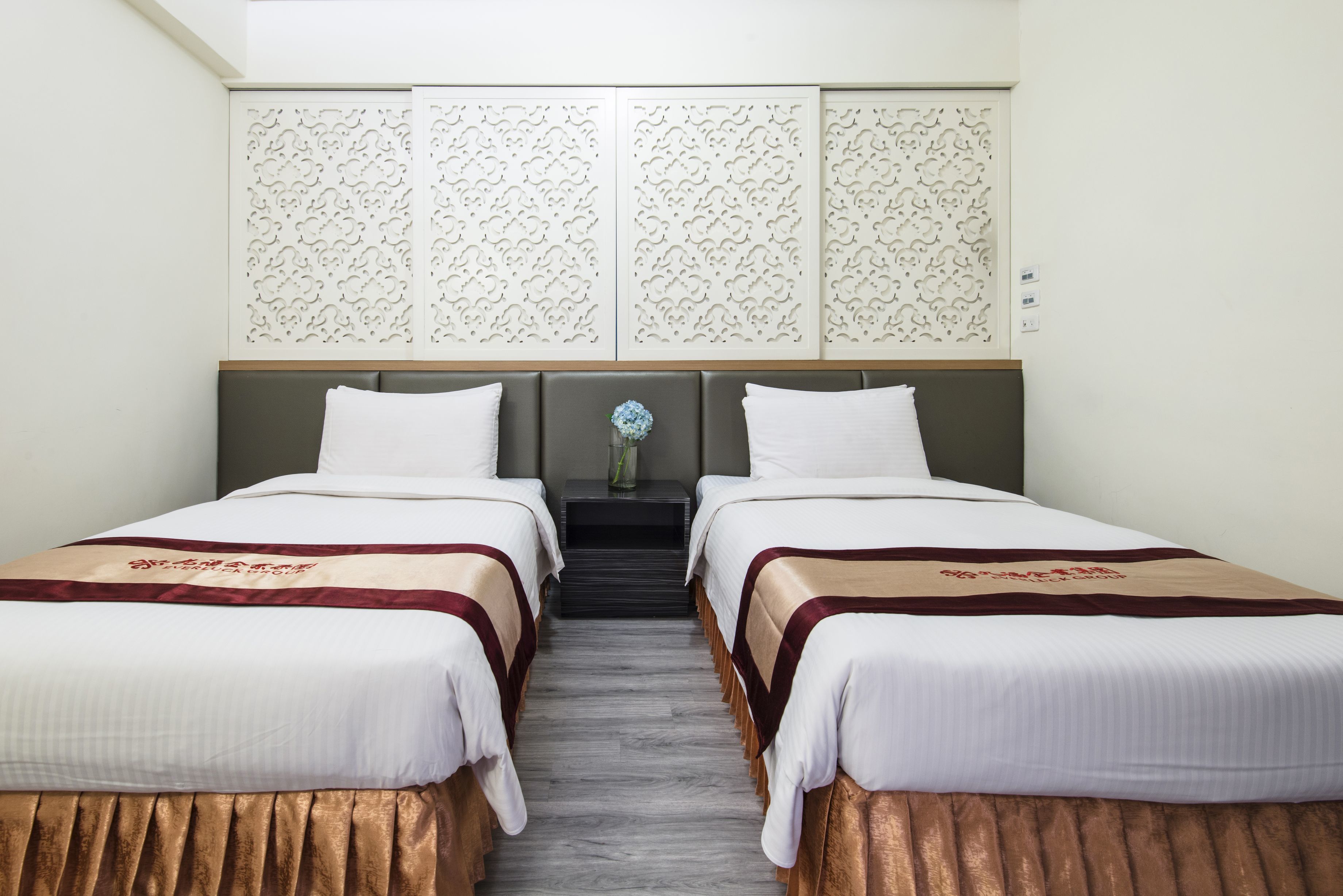 Una habitación de hotel con dos camas, un cabecero decorativo, una mesita con un florero y suelo de madera.