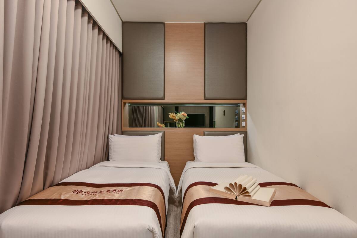 Une chambre d’hôtel avec deux lits, une tête de lit en bois, une petite fenêtre avec un vase et un miroir.