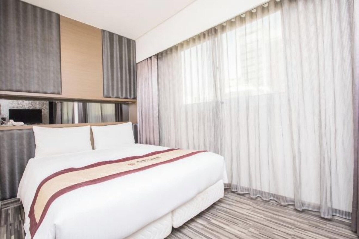 A modern hotel room with a large bed, a flat-screen TV, and sheer curtains.