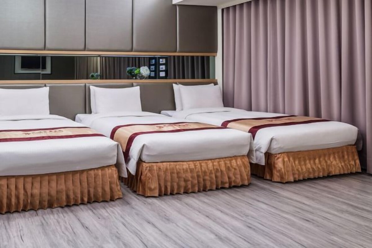 A hotel room with three beds, each with a white mattress and a brown skirt. There is a wooden headboard with a mirror and shelves above the beds.