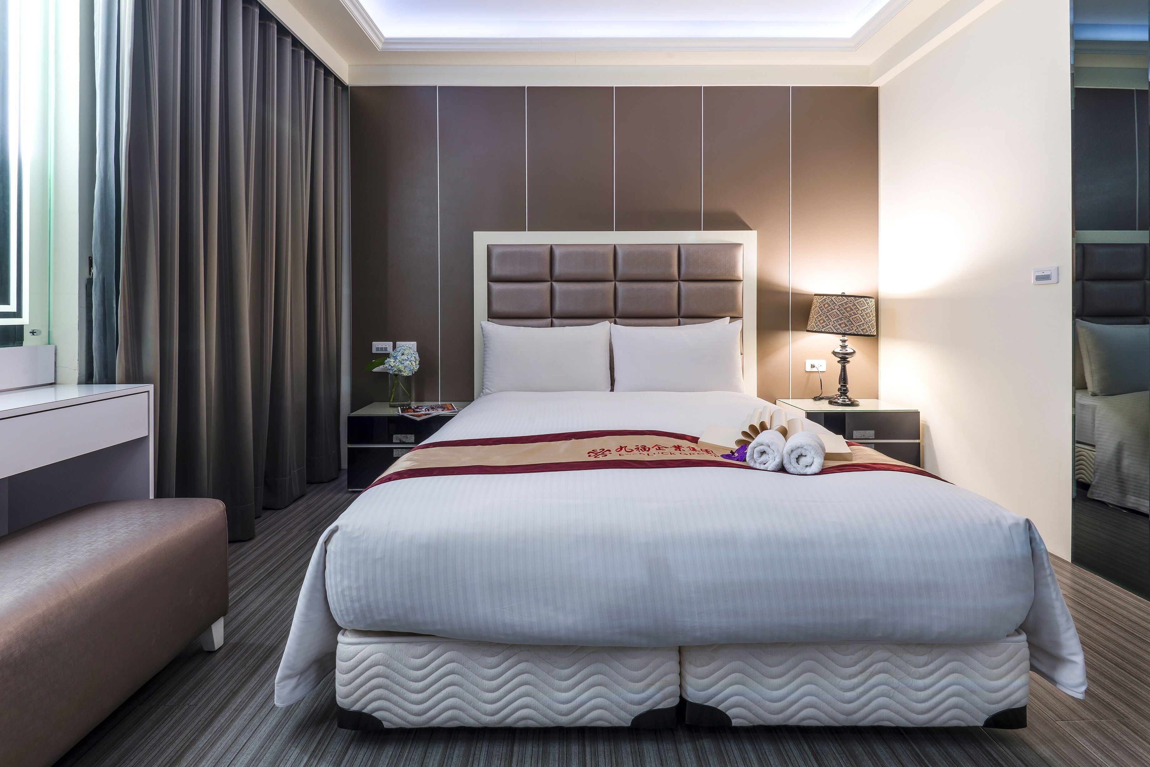 A hotel room with a large bed, a bedside table with a lamp, a nightstand, and a bench.