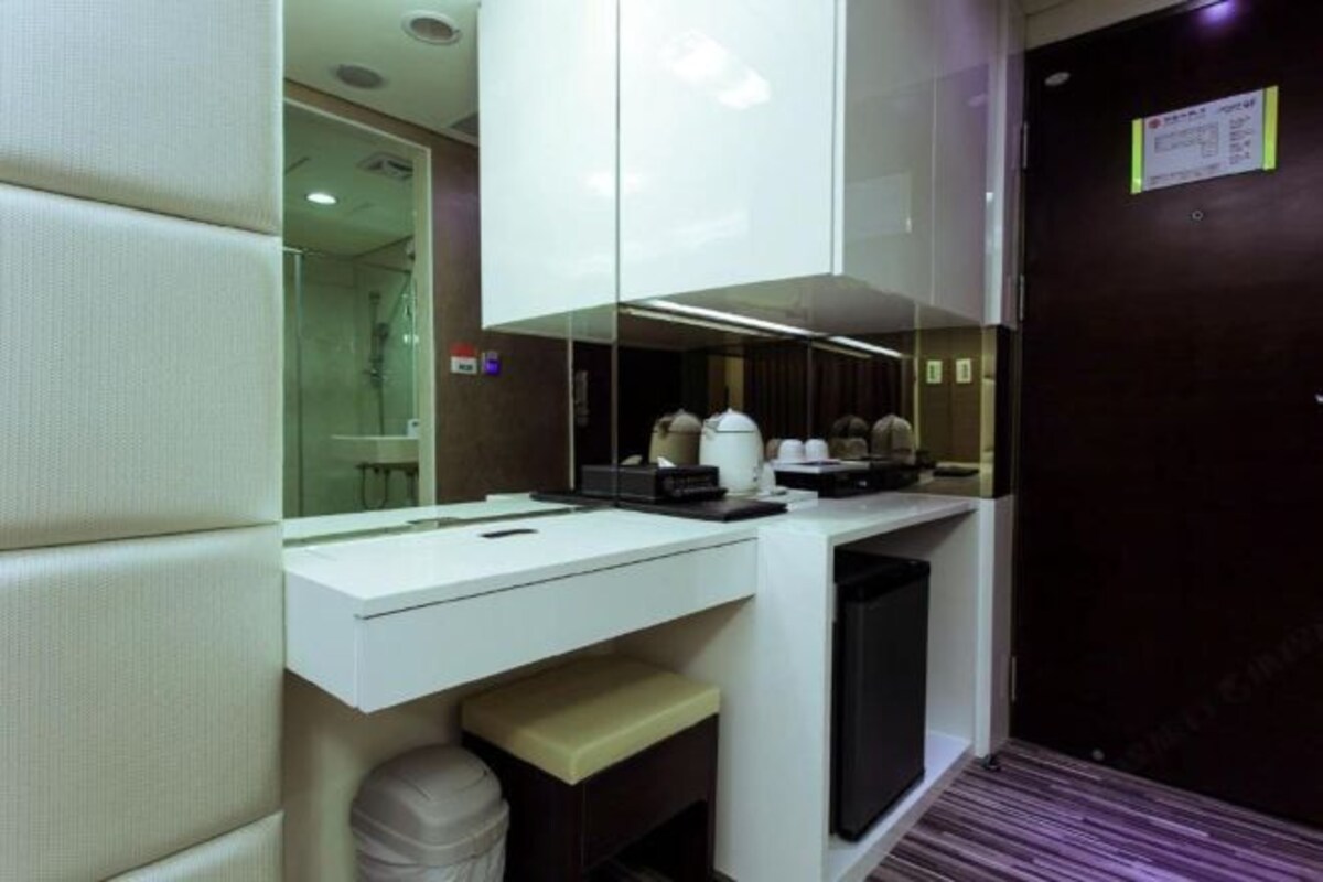 A compact hotel room with a white desk, a small trash bin, and a dark-colored door.