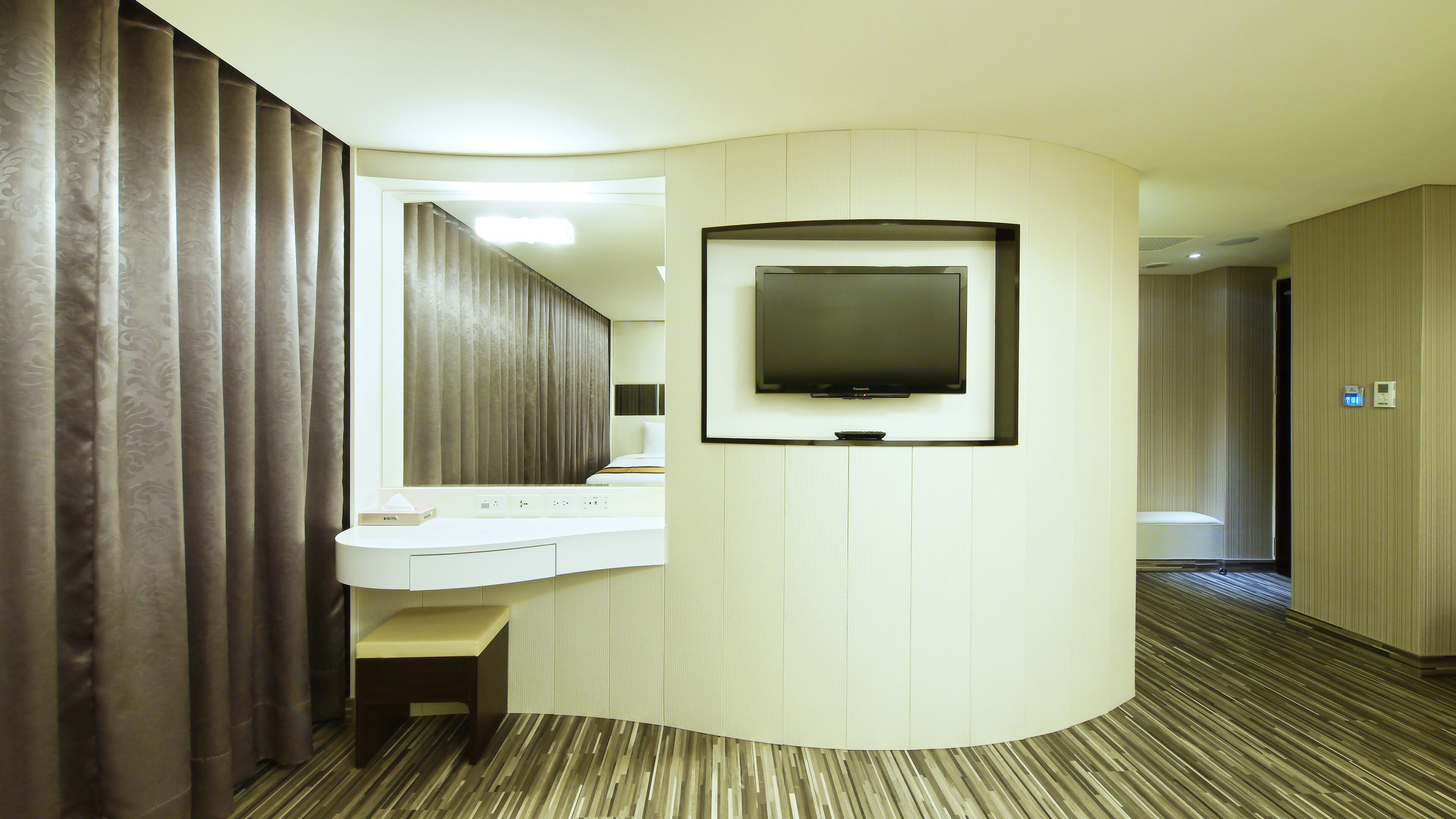Habitación de hotel con tocador curvo, televisor de pantalla plana montado en la pared, suelo de madera y un espejo grande.