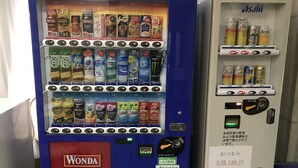 Een automaat met diverse drankjes en snacks, waaronder blikjes Wonda en Asahi, en een pinautomaat.