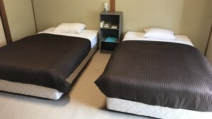 Twee enkele bedden met witte lakens en donkere hoofdborden, een klein nachtkastje met een lamp, en een wandplank met toiletartikelen.