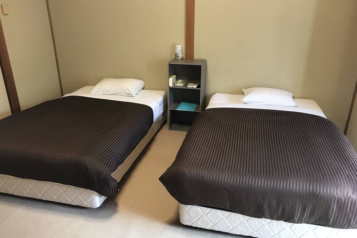 Two single beds with white linens and dark headboards, a small bedside table with a lamp, and a wall-mounted shelf with toiletries.