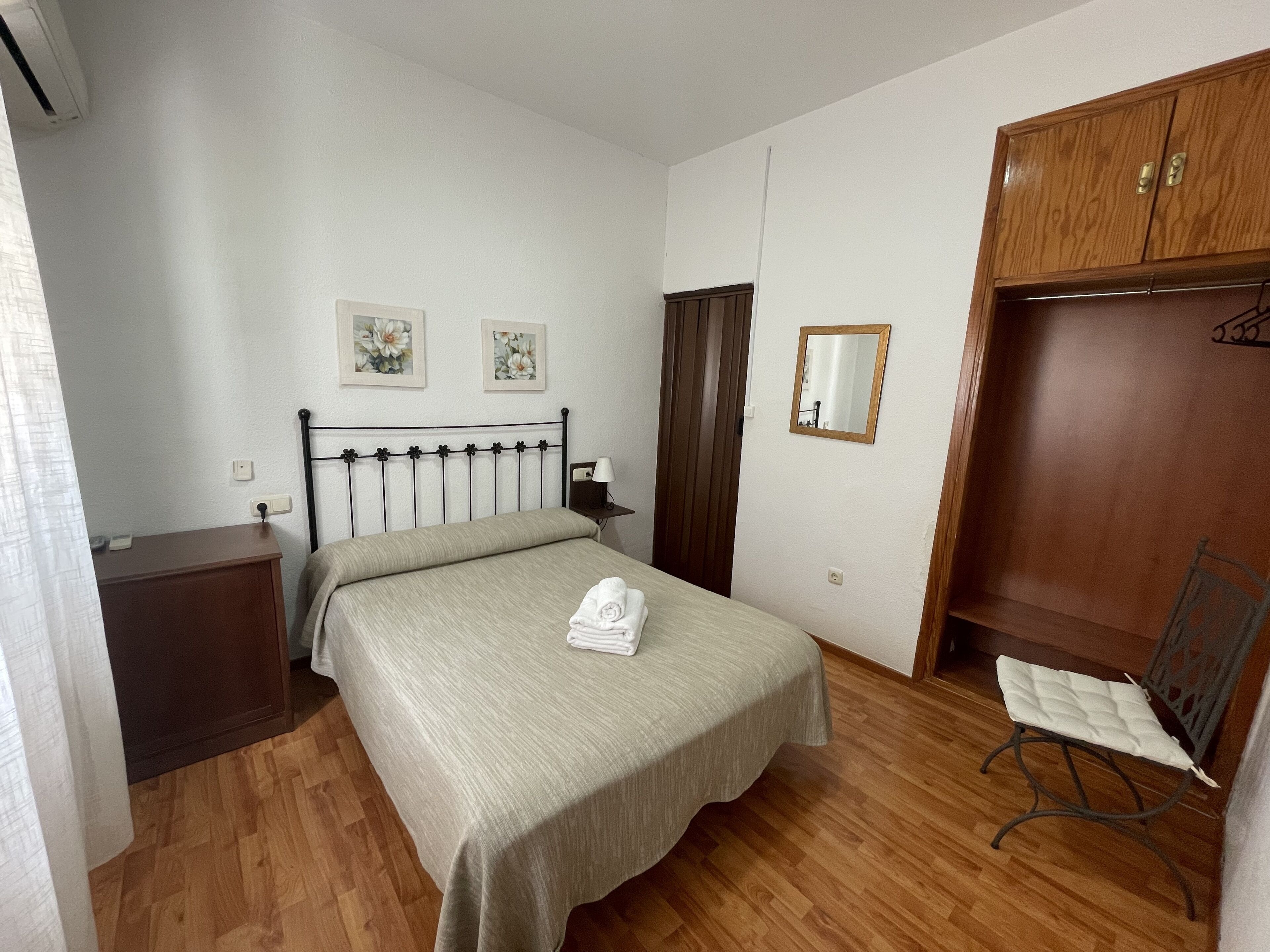 Un dormitorio con una cama, un armario de madera, una silla, una mesita de noche, un espejo y dos cuadros florales.