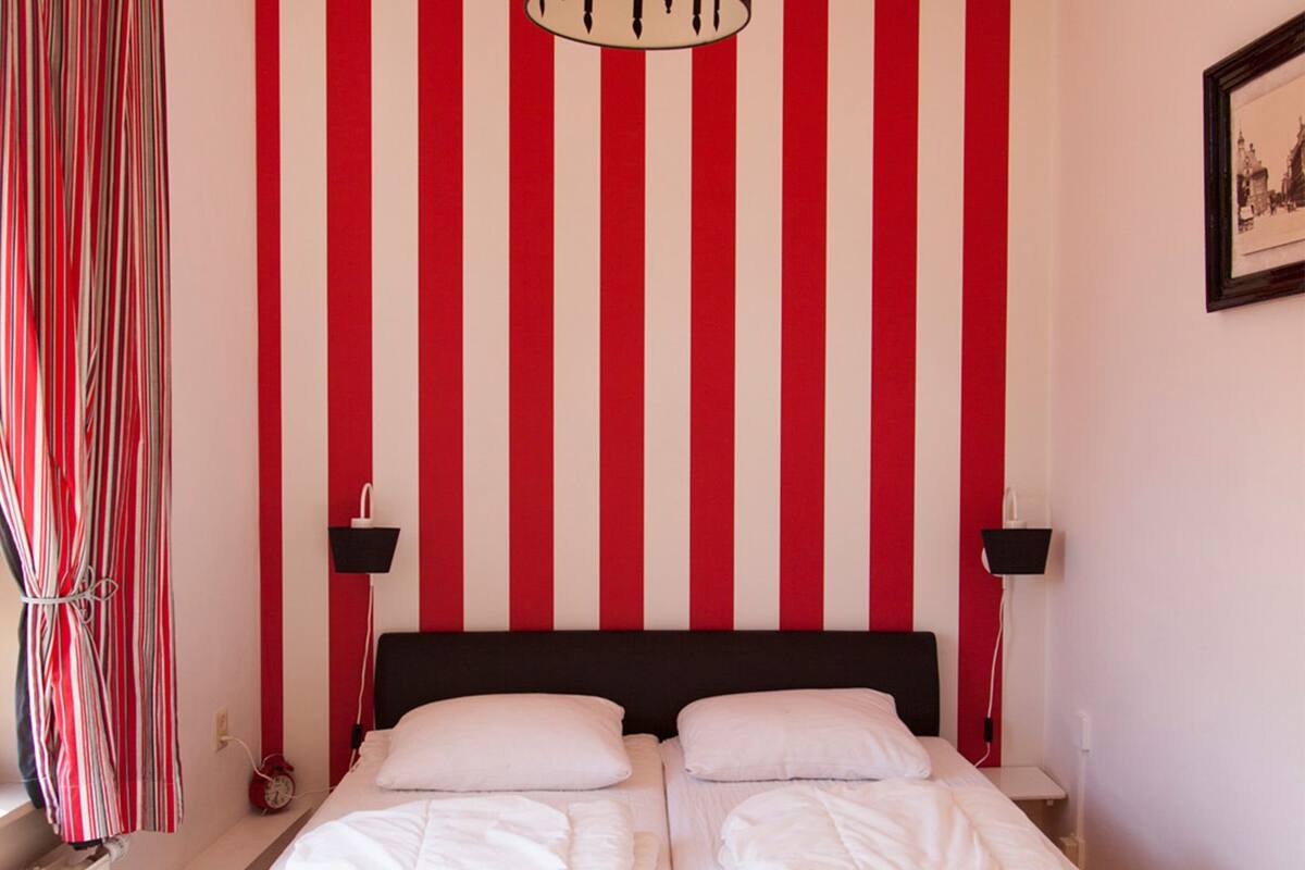 Een slaapkamer met een gestreepte rode en witte muur, een zwart hoofdeinde, twee nachtkastjes met lampen en een raam met gordijnen.
