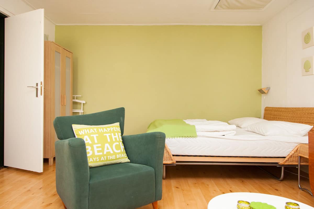 Een kamer met een groene fauteuil, een bed met witte beddengoed, een klein rond tafeltje met groene objecten en een houten vloer.