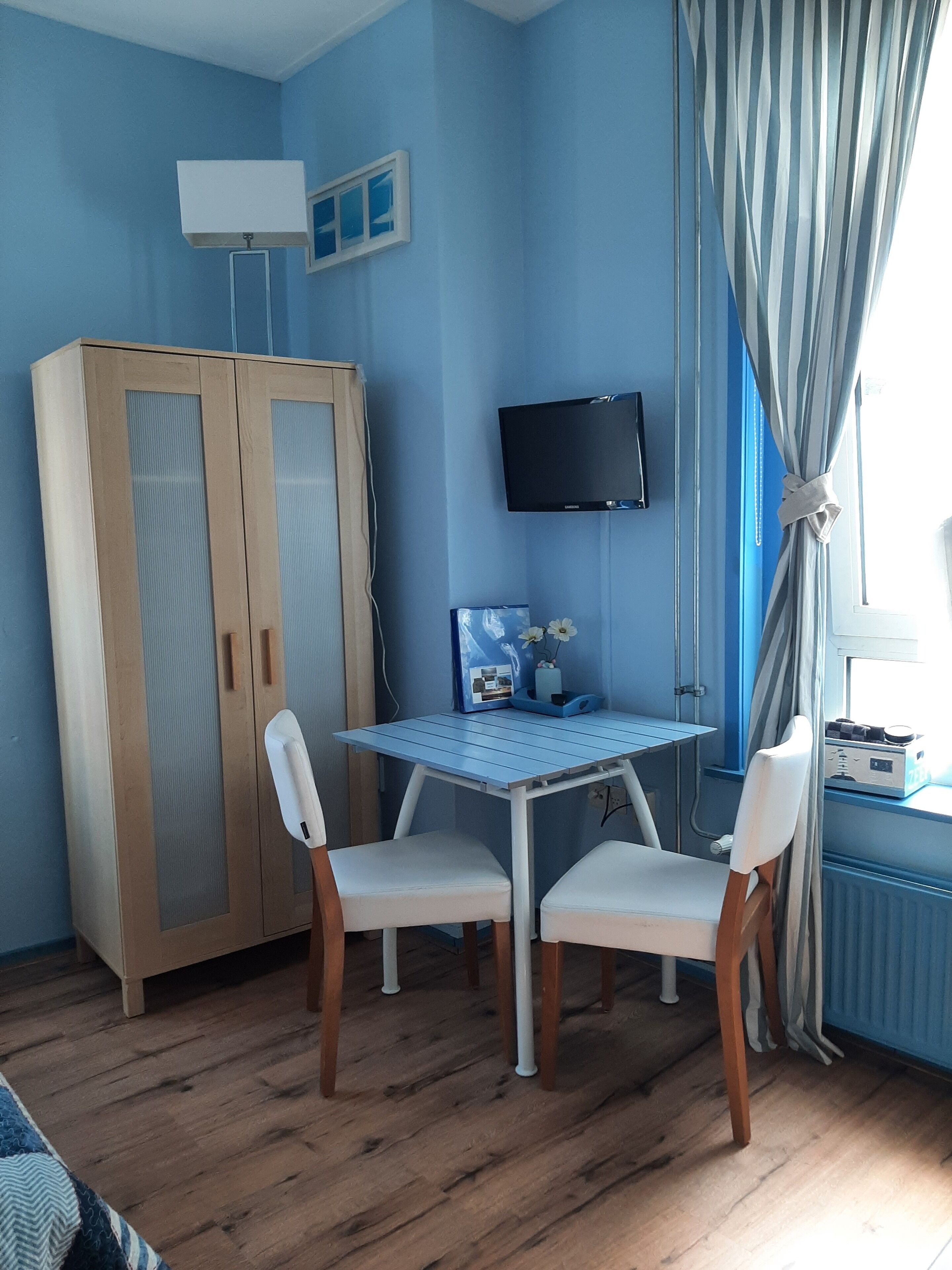 A room with a wooden wardrobe, a TV mounted on the wall, a table with two chairs, and a window with curtains.