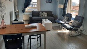 Un salon moderne comprenant un canapé, deux fauteuils, une table basse et une table à manger avec des chaises. Il y a deux fenêtres avec des rideaux, un radiateur et un tableau accroché au mur.