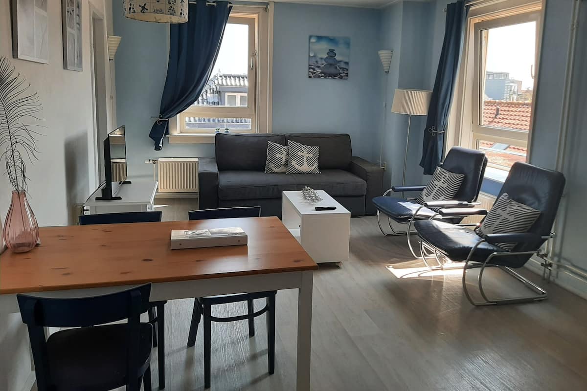 Een moderne woonkamer met een bank, twee fauteuils, een salontafel en een eettafel met stoelen. Er zijn twee ramen met gordijnen, een radiator en een schilderij aan de muur.