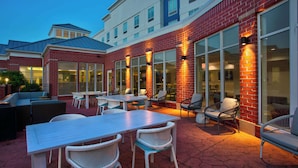 An outdoor patio area with a long table and several chairs, surrounded by brick walls and large windows.