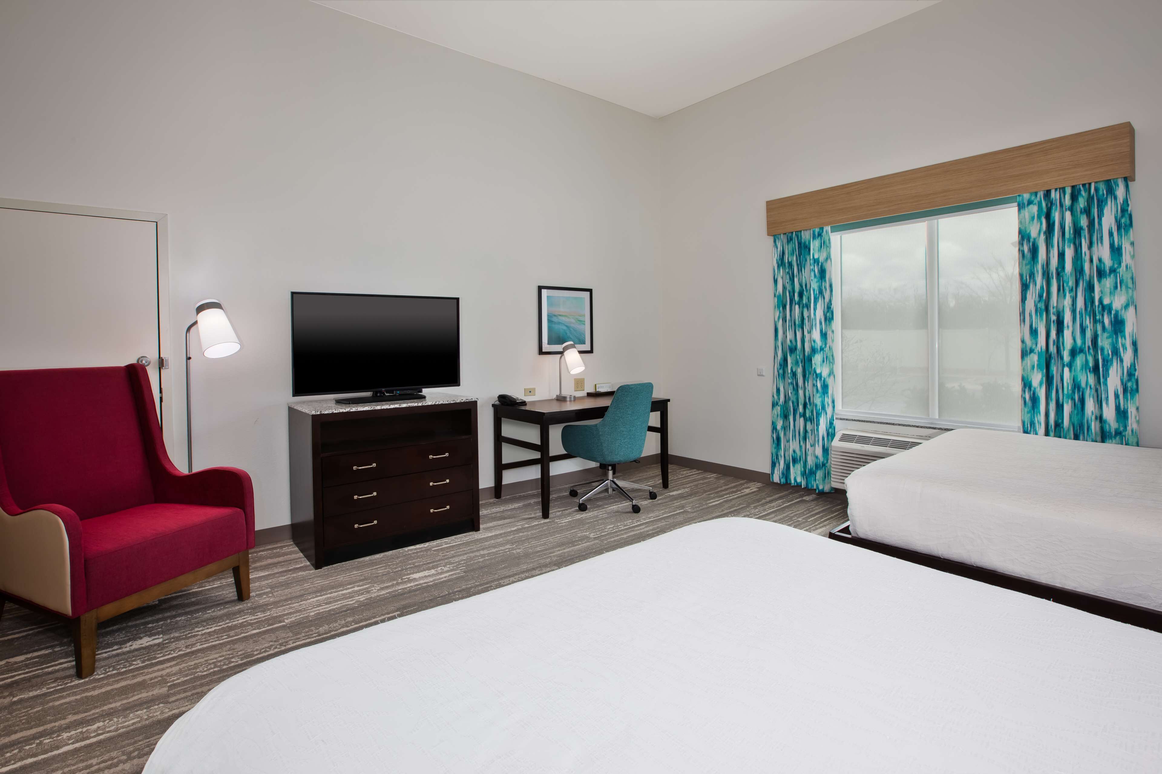 A hotel room with a bed, a desk with a chair, a TV, a red armchair, and a window with curtains.