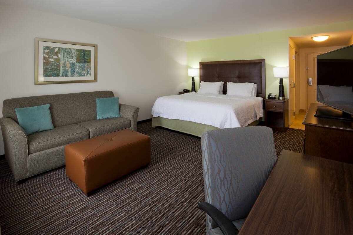A hotel room with a bed, a sofa, an ottoman, a desk, and a television.