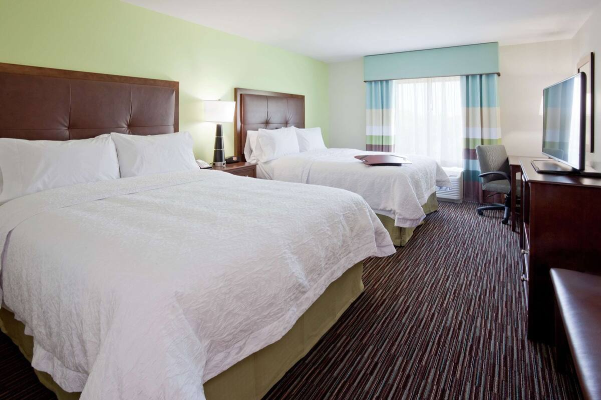 A hotel room with two beds, a desk, a chair, a TV, and a window with curtains.