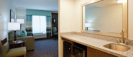 Habitación de hotel con kitchenette, un espejo grande, un sofá, una silla y vistas al vestíbulo del hotel.