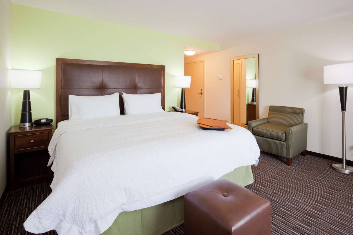 A hotel room with a large bed, a bedside table with a lamp, a chair, and a mirror.