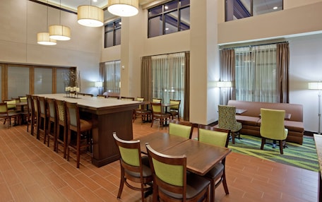 A spacious hotel dining area with a long wooden table, multiple chairs, and a bar area. There are large windows with curtains and a high ceiling with hanging lights.