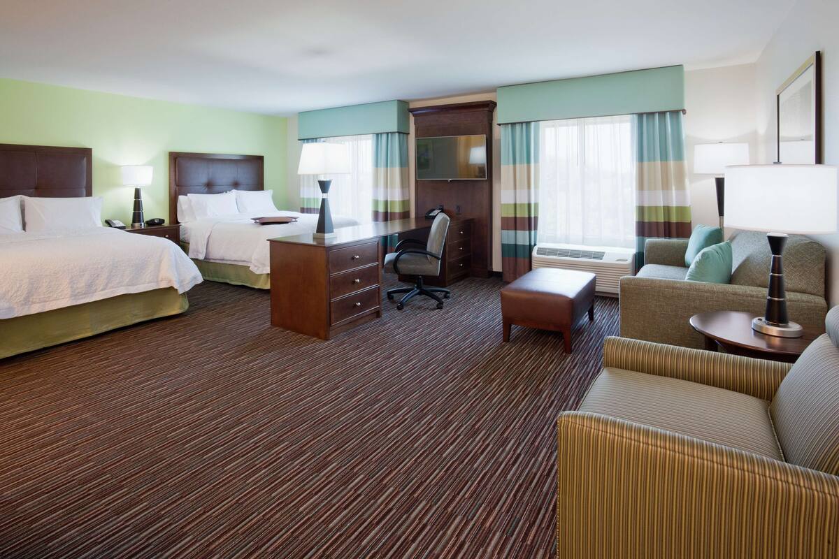 A hotel room with two beds, a desk, a chair, a sofa, and a large window with curtains.