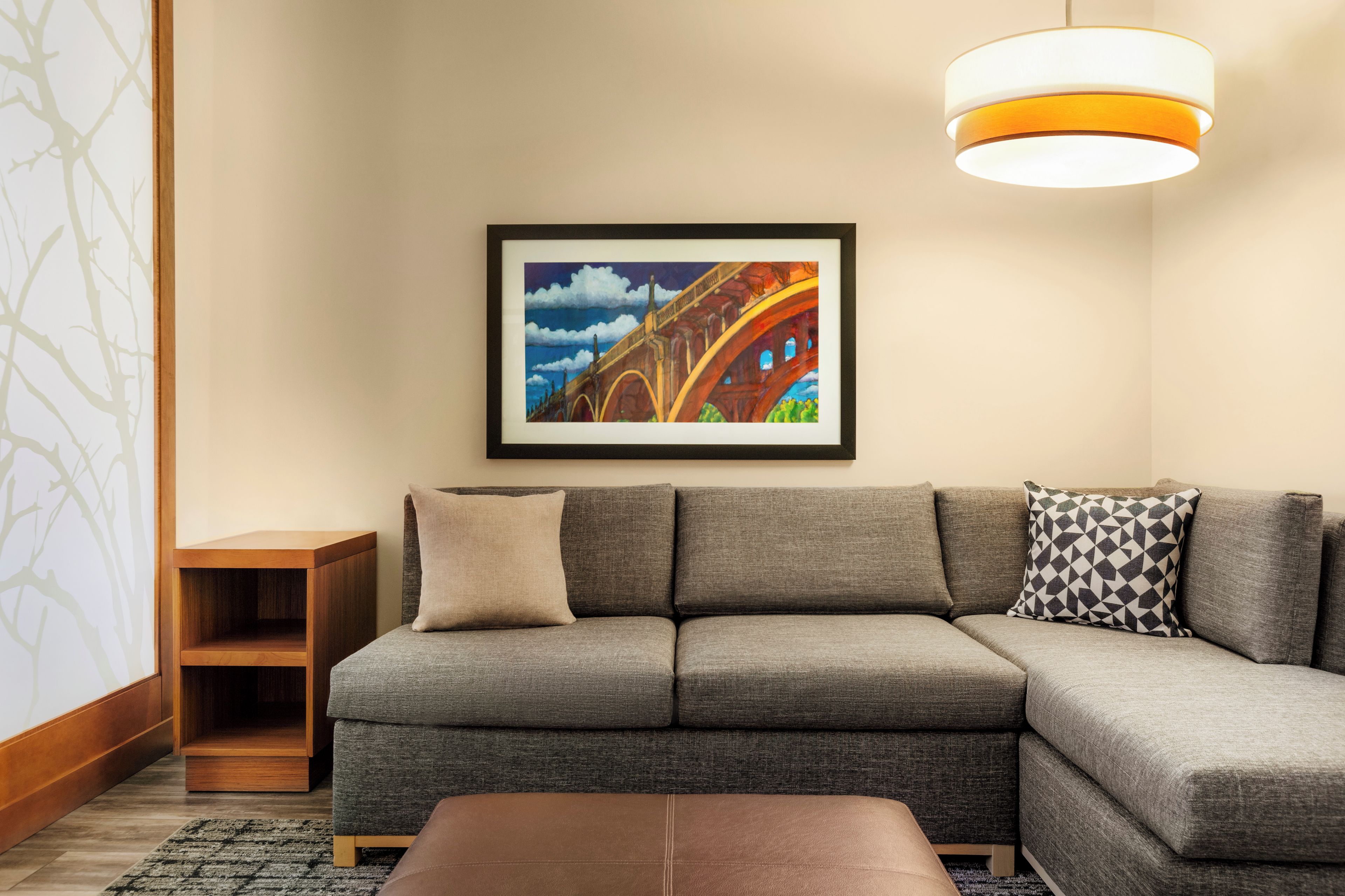A modern living room with a sectional sofa, a small wooden side table, and a framed painting of a bridge.