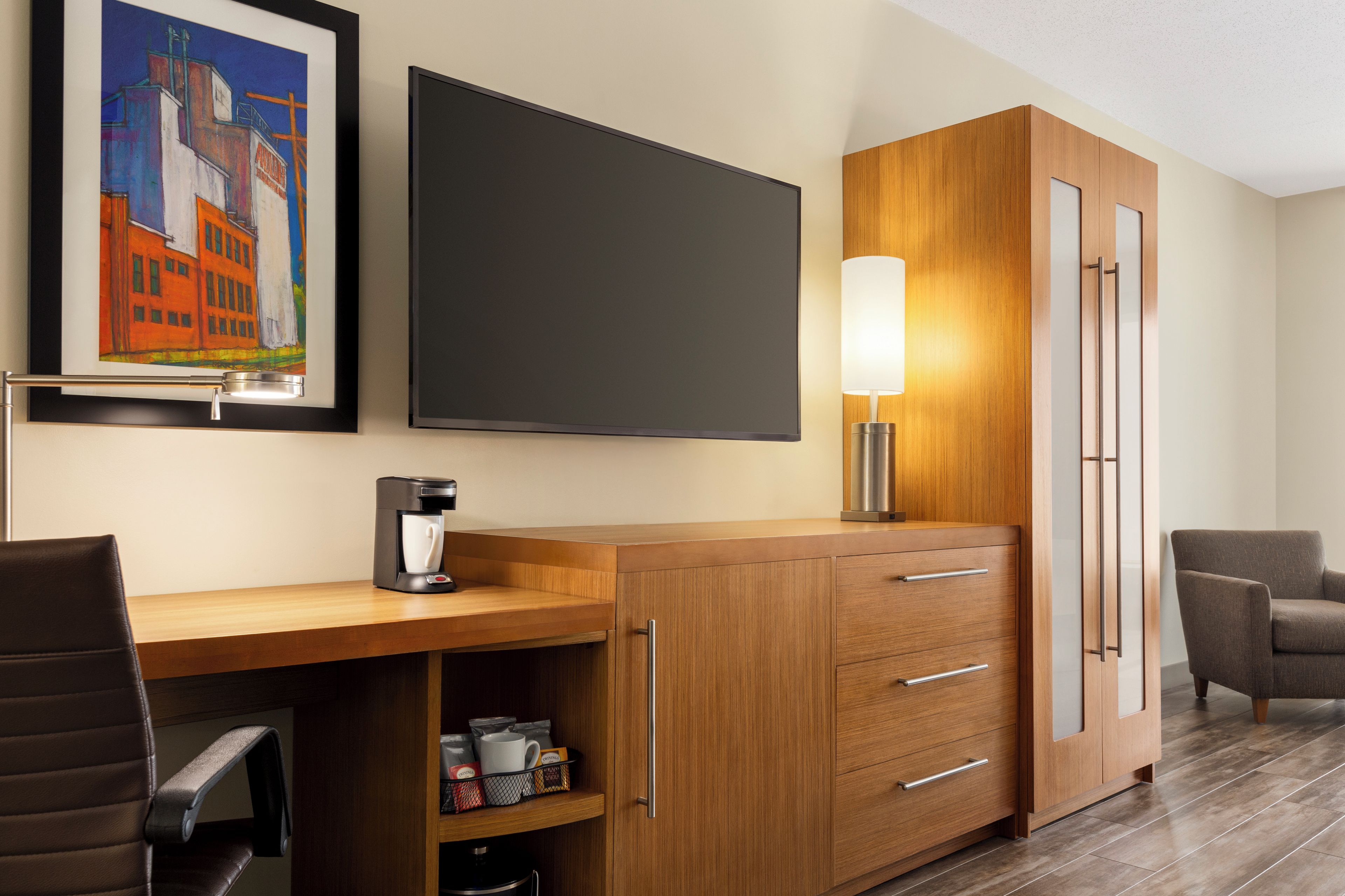 A hotel room with a wooden desk, a coffee maker, a chair, a cabinet with drawers, a framed picture on the wall, and a flat-screen TV.